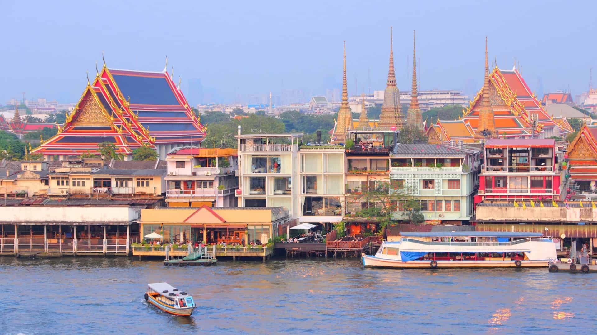 Colorful riverside buildings and boats with ornate temple rooftops in Bangkok, Thailand, under a clear sky.