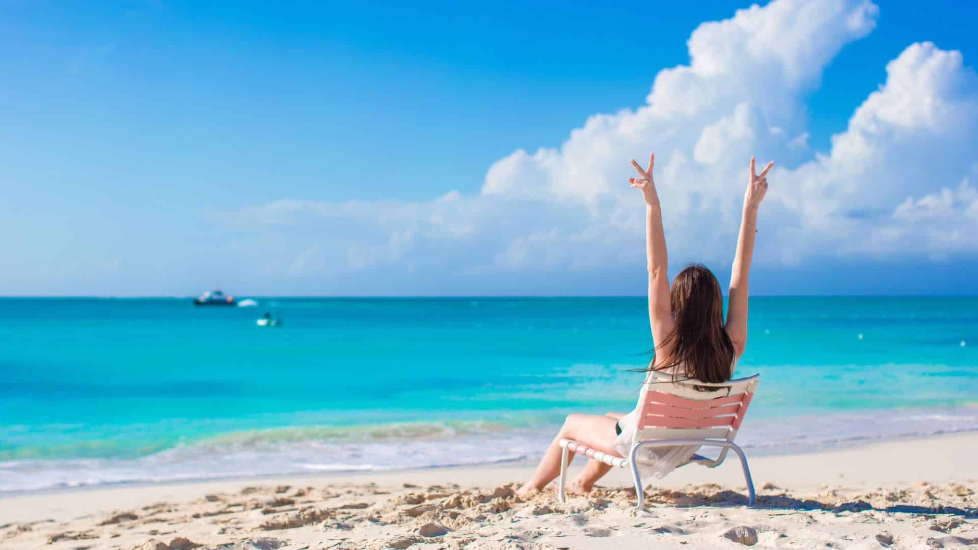 Woman sitting on a beach chair facing the sea, raising both arms in a peace sign toward the blue sky.