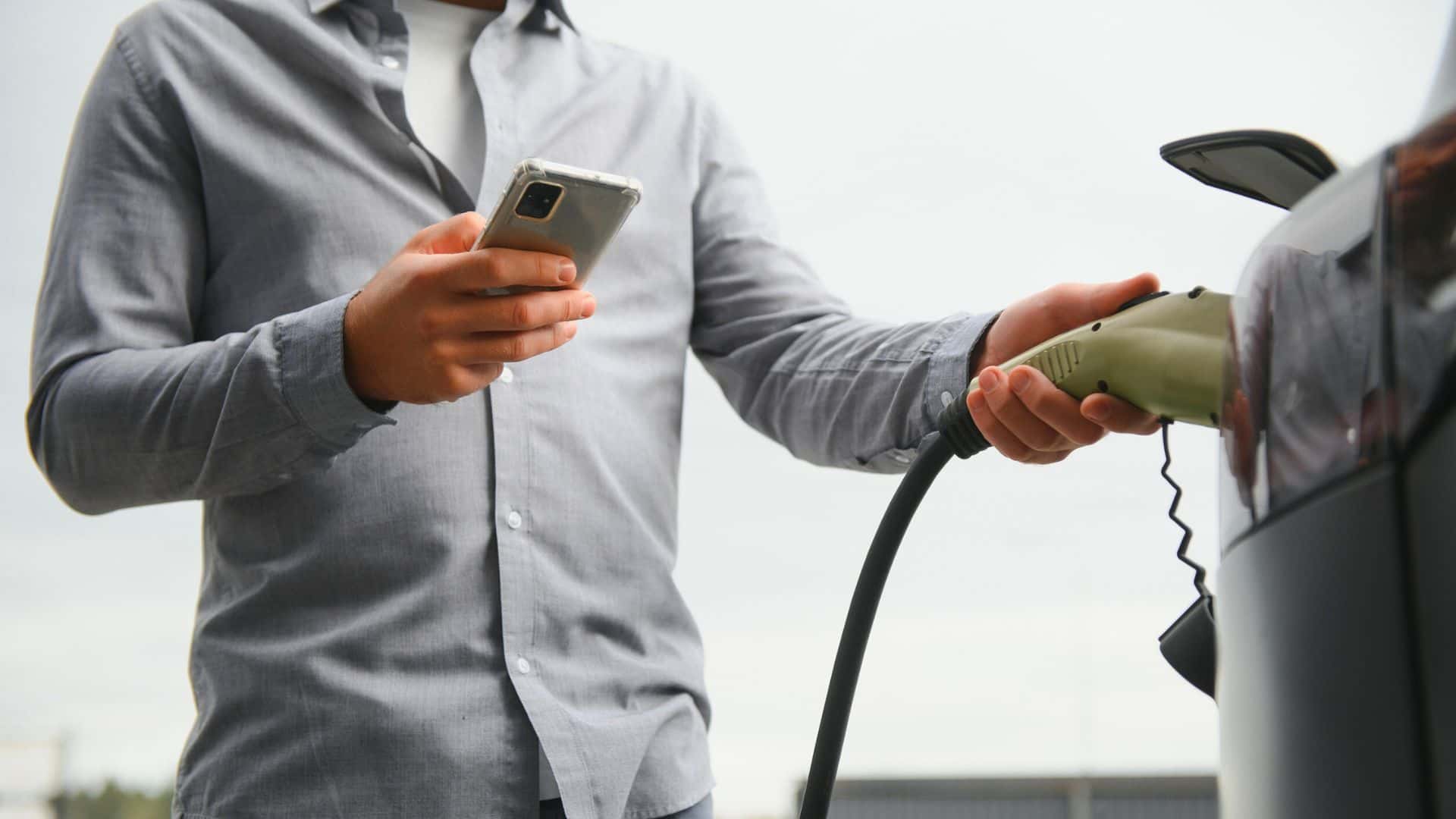 A person charging an electric car while holding a smartphone.