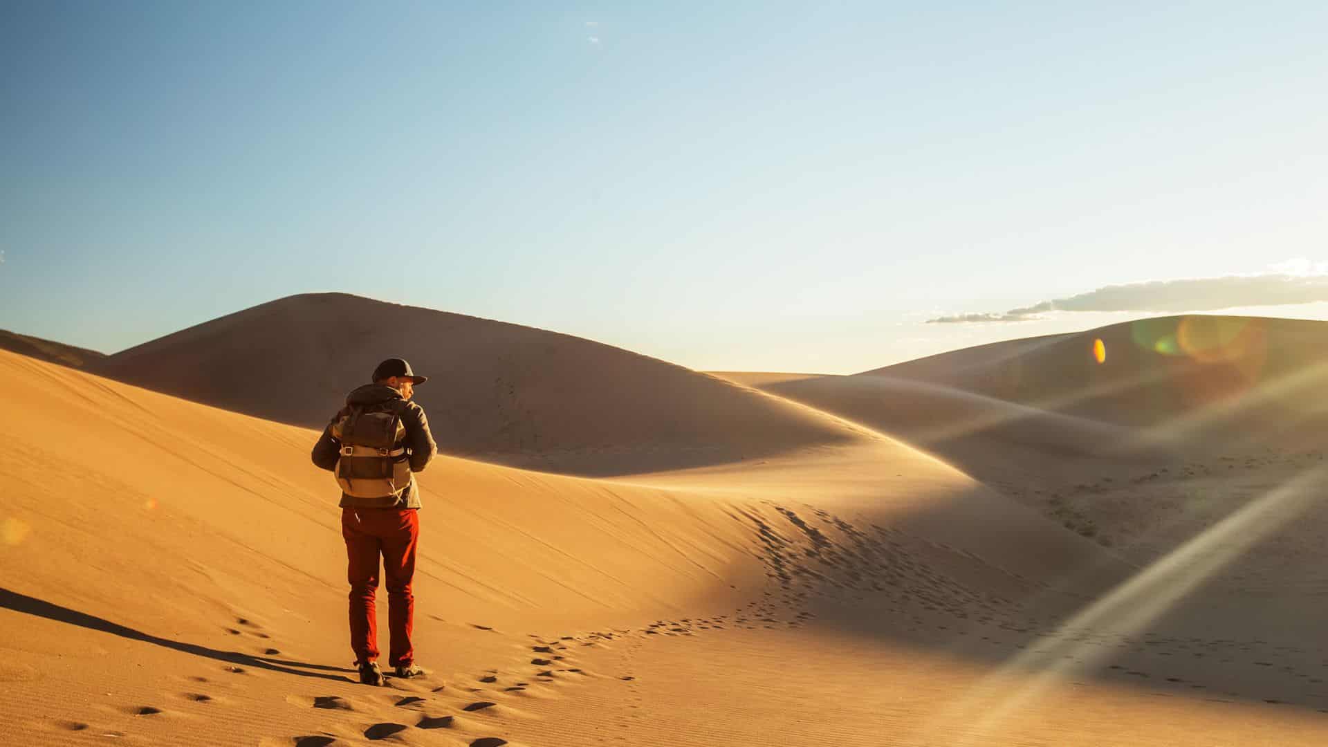 A person with a backpack walks alone across sunlit sand dunes in a desert landscape.