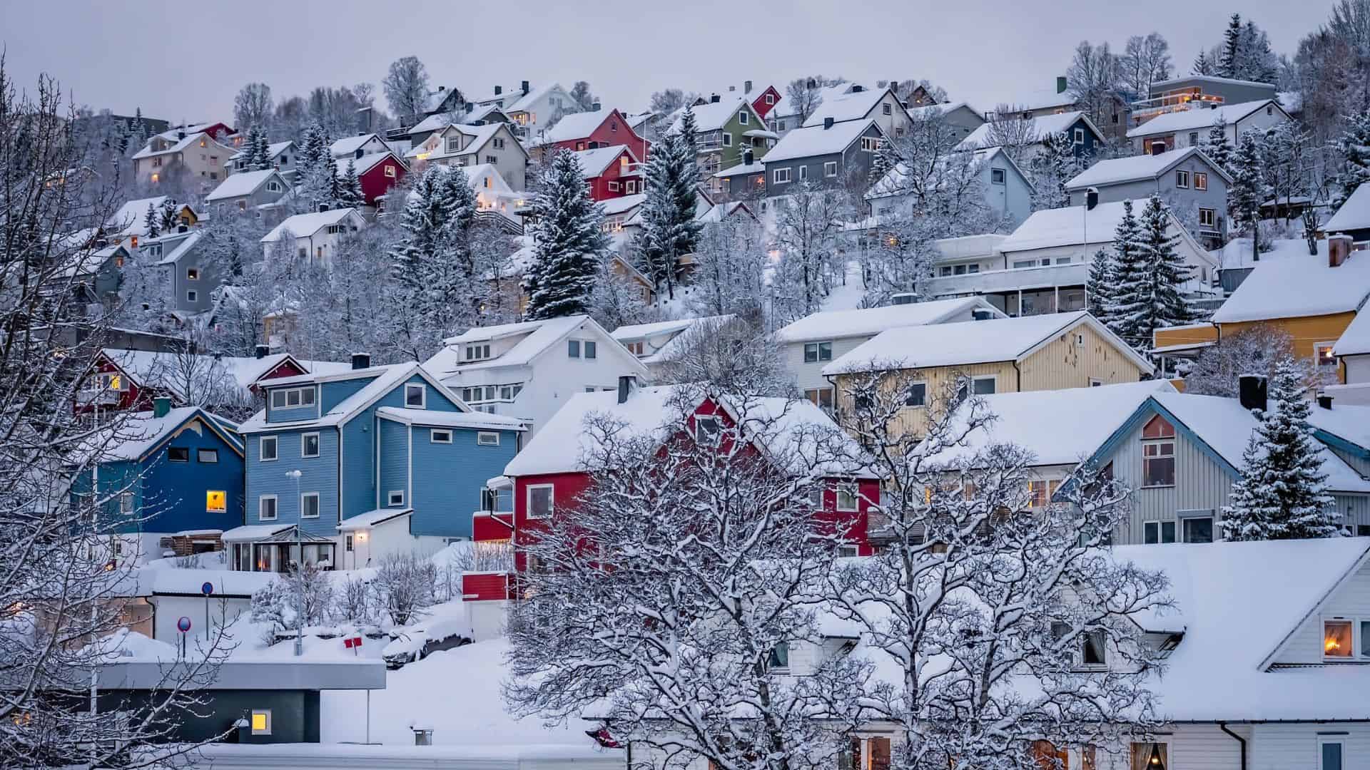 Snow-covered houses in a hilly neighborhood with trees in winter, featuring colorful homes and a calm atmosphere.