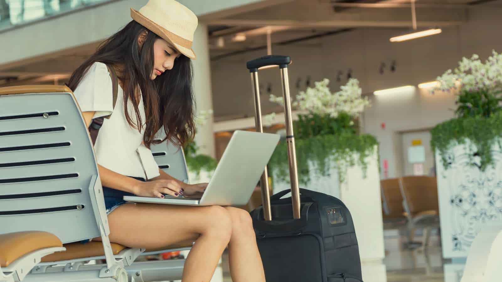 Woman in a hat using a laptop while sitting in an airport with a suitcase beside her.