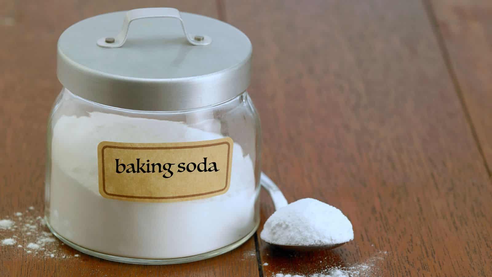 A jar labeled "baking soda" sits on a wooden table with a spoonful of baking soda beside it.