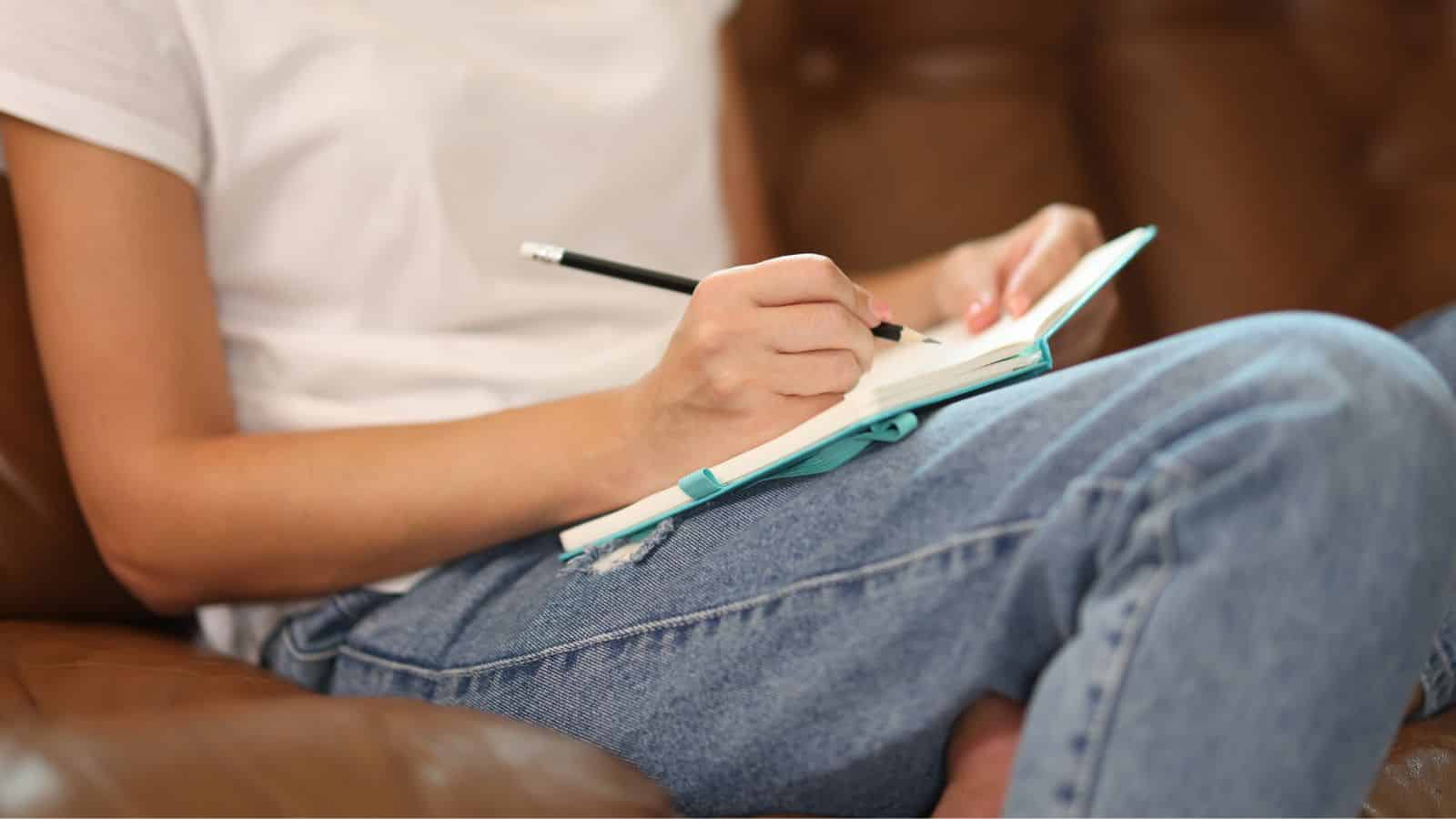 A person in jeans and a white shirt writes in a notebook while sitting on a brown sofa.