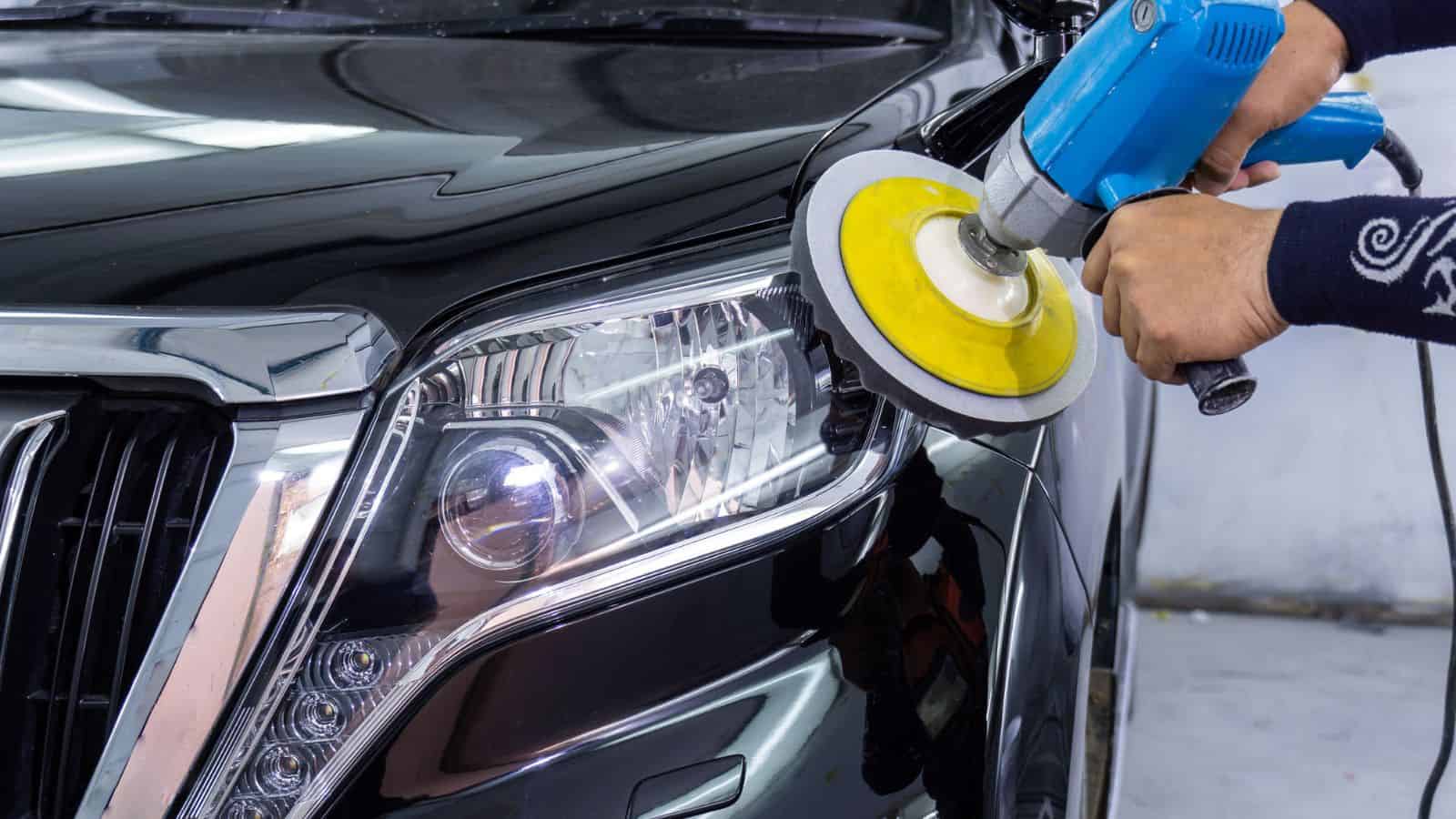A person polishing the headlight and hood of a black car with a power buffer.