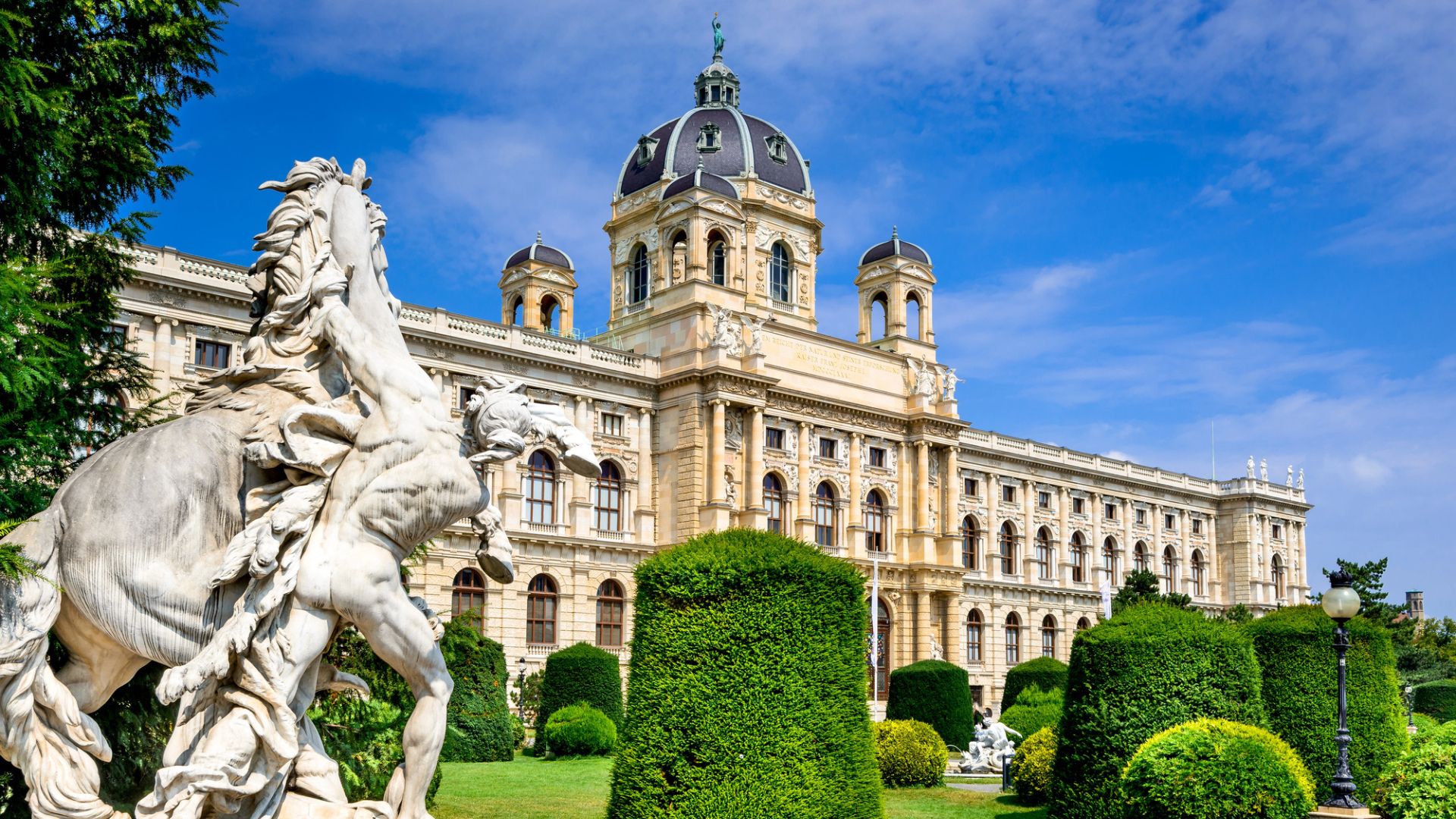 Baroque building with domes, trimmed hedges, and a horse statue in a sunny garden under a blue sky.