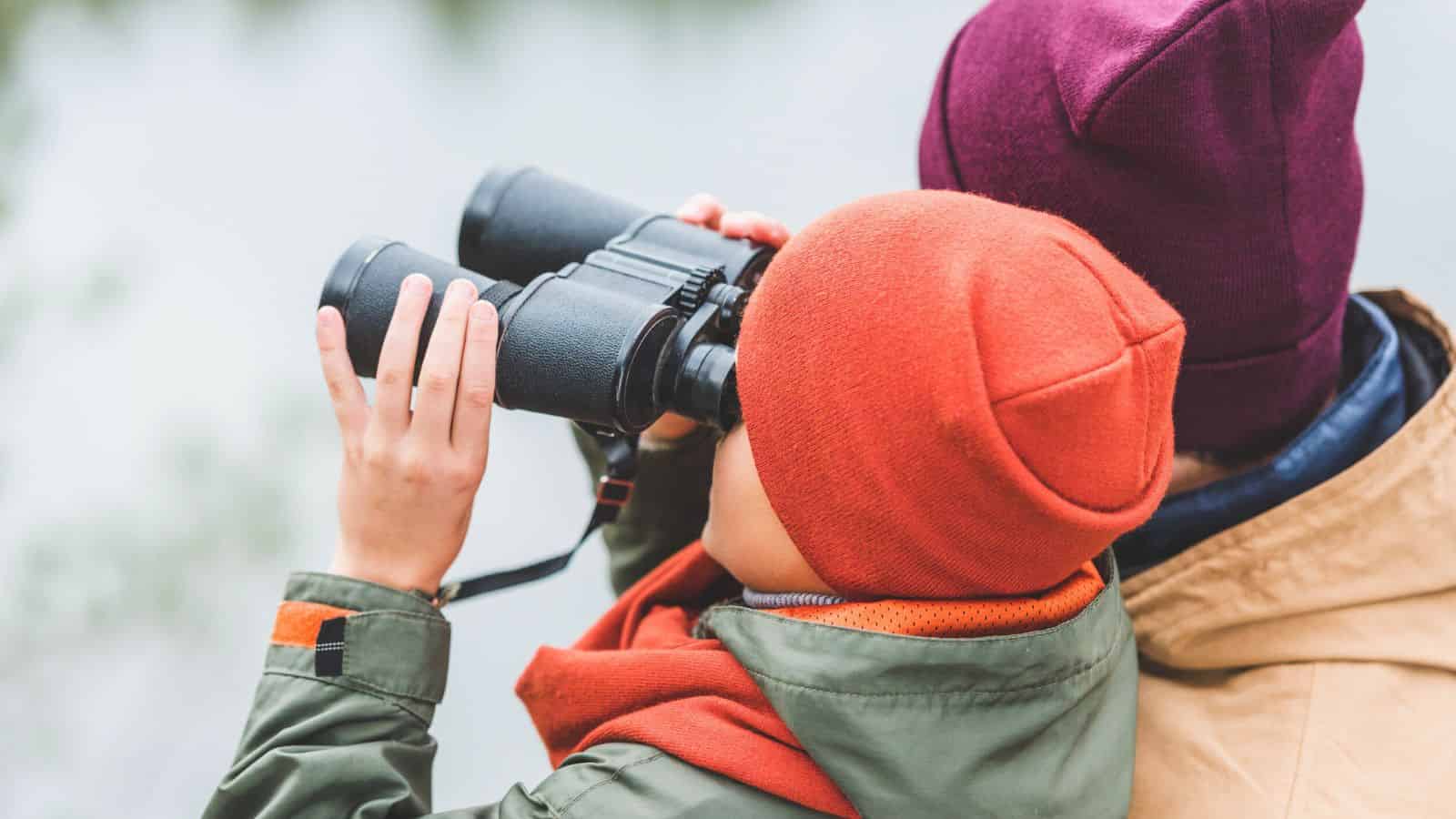 Two people in jackets and beanies look through binoculars outdoors, facing away from the camera.