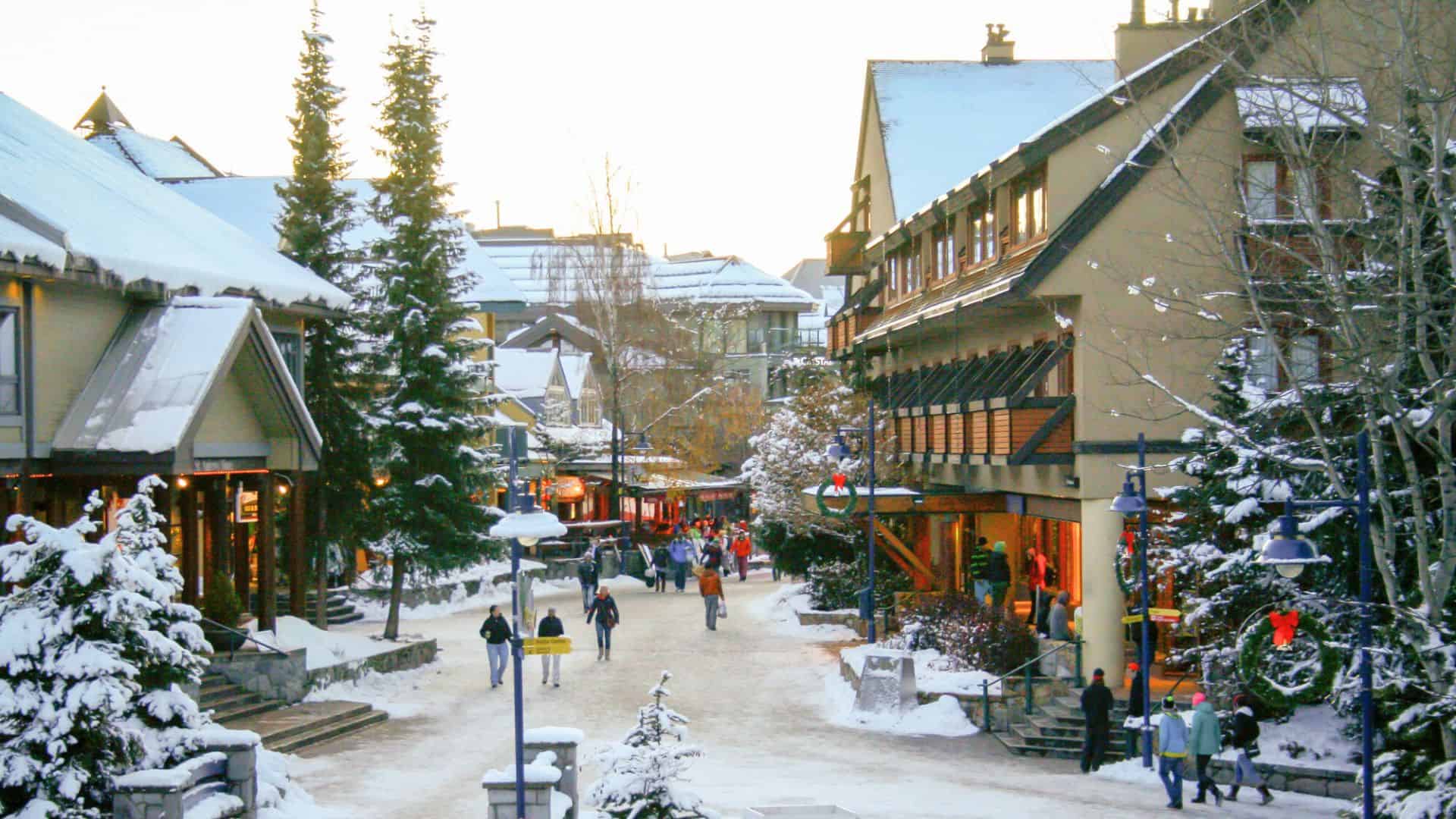 Snowy pedestrian street lined with shops and trees, people walking, and warm lights glowing from buildings.