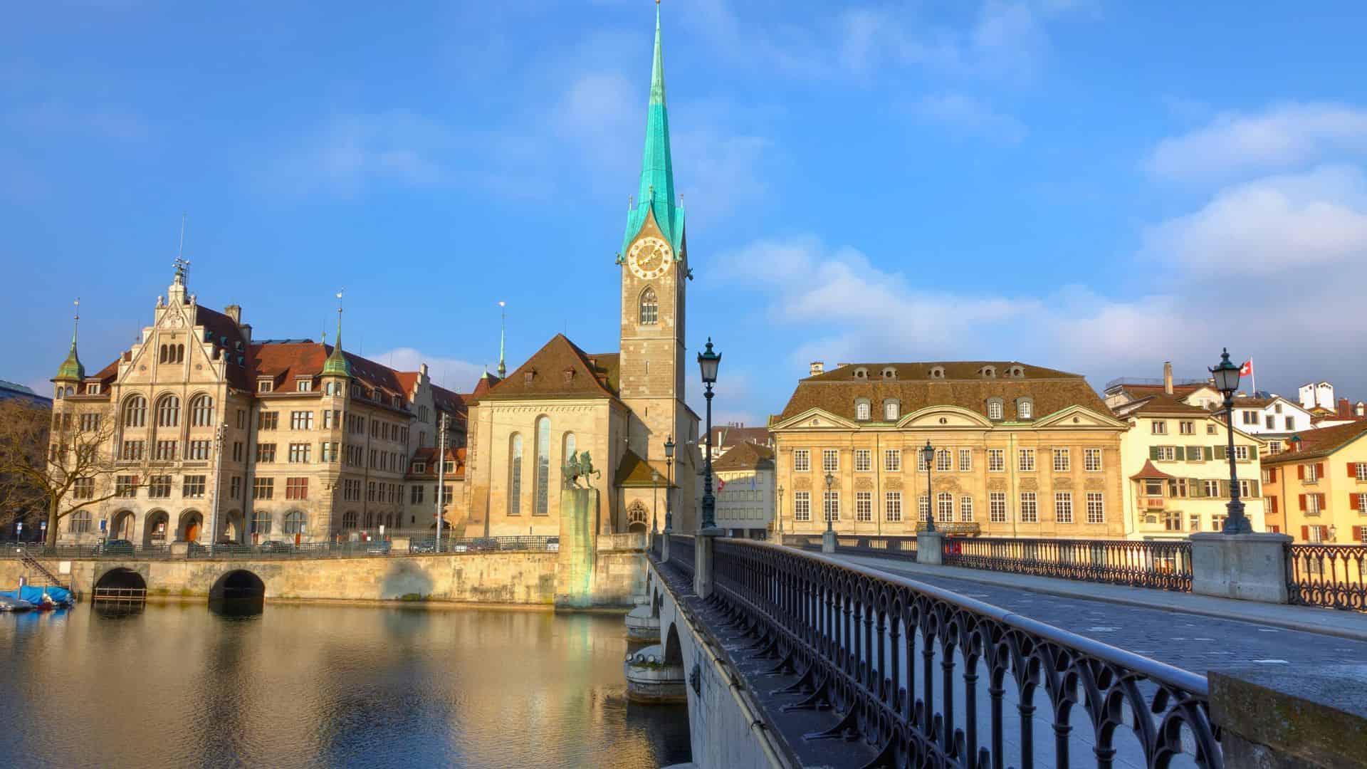 Historic buildings and a church with a turquoise spire by the river in Zurich, Switzerland, under a blue sky.
