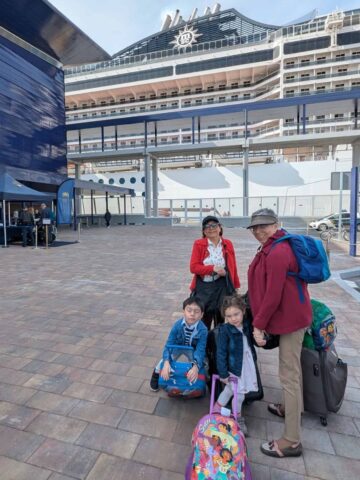 Four people with luggage stand outside a cruise ship terminal, ready to embark, with a large cruise ship in the background—an ideal scene for sharing first time cruise tips.
