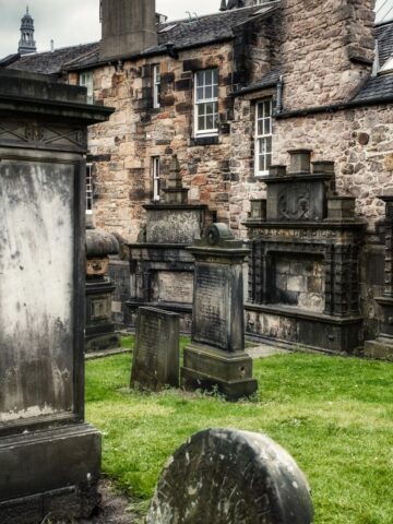 Old gravestones and tombs stand on grass near a stone building with multiple windows, evoking the eerie charm found in haunted places in Europe.