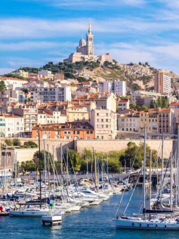 Boats docked in a marina with Marseille’s hillside cityscape and Notre-Dame de la Garde basilica in the background—a perfect scene for a one day trip Marseille experience.