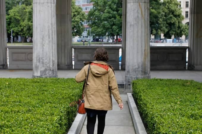 A person in a tan coat walks between hedges and stone columns toward an open area outside.