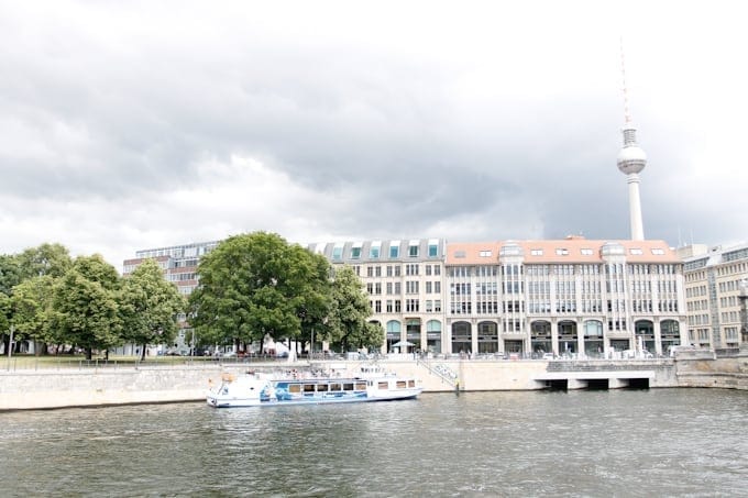 A tour boat on a river with buildings, trees, and the Berlin TV Tower in the background.