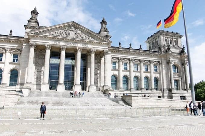 The Reichstag building in Berlin with a large German flag and people outside on a sunny day.