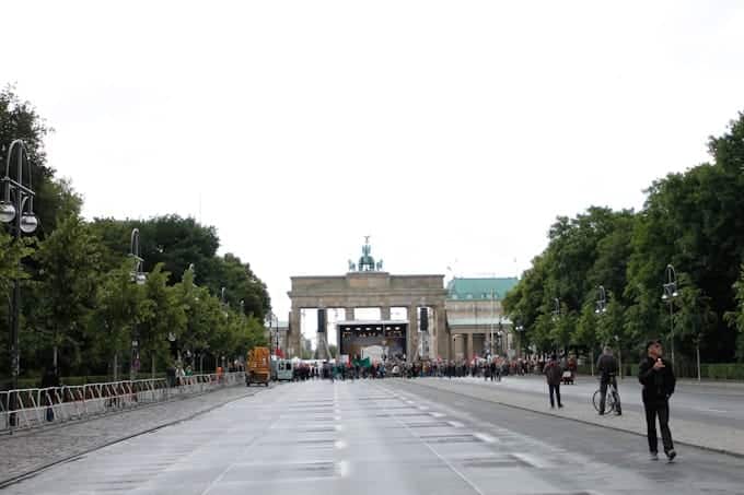 Crowd gathers near the Brandenburg Gate on a wet, tree-lined street in Berlin, with cloudy skies above.