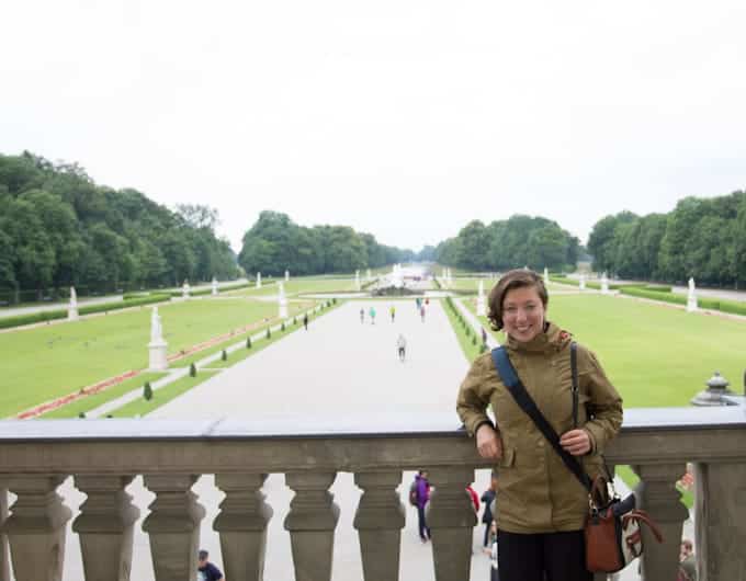 Person in a green jacket standing on a stone balcony overlooking a formal garden with lawns and statues.