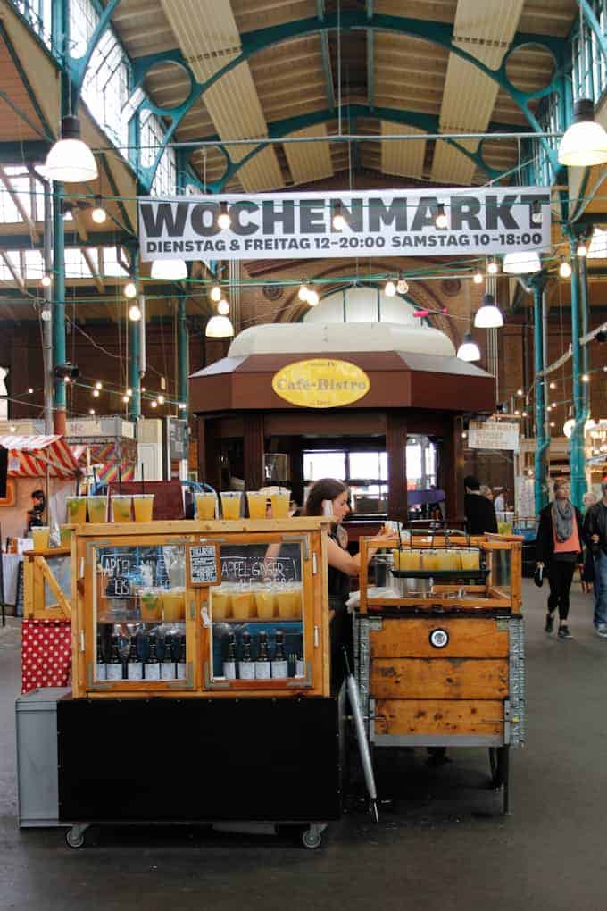 Indoor market with juice stands and a large "Wochenmarkt!" sign hanging above the stalls.