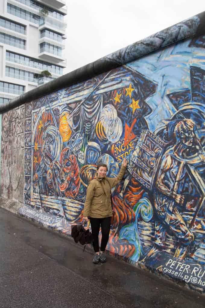 A woman stands by a colorful mural on the Berlin Wall, pointing at the artwork on a cloudy day.