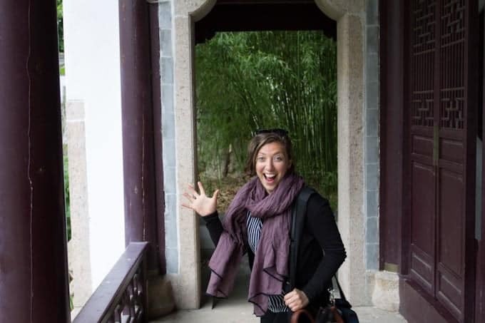 Smiling woman with a purple scarf waves while standing in a corridor with bamboo in the background.
