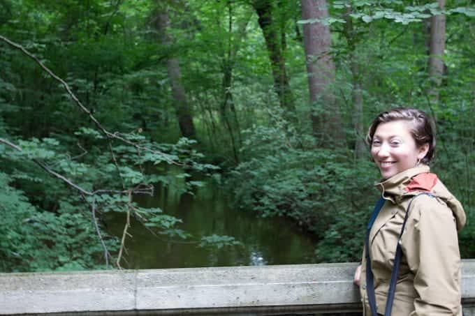Woman in a brown jacket smiles on a bridge in a lush green forest by a creek.
