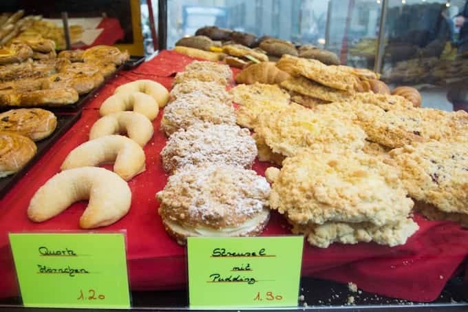 Assorted German pastries displayed on trays with handwritten price signs in a bakery.