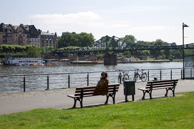 A person sits alone on a bench by a river, with a bridge and buildings in the background.