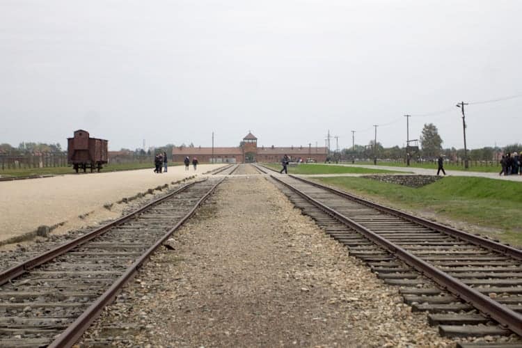 Railway tracks lead to a large brick building under a cloudy sky, with people walking nearby, evoking the experience of visiting Poland.