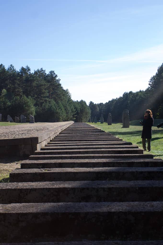 Wide stone steps lead toward a grassy area with tall stones; a person stands to the right under a blue sky, evoking the experience of visiting Poland’s historic landmarks.