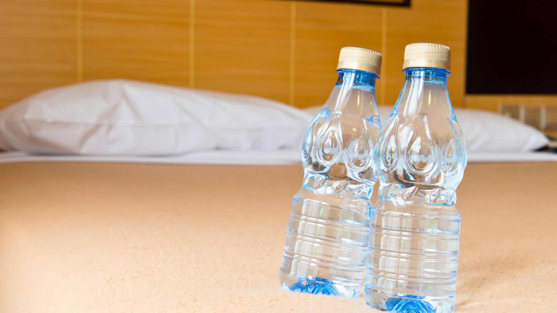 Two plastic water bottles on a beige bedspread with pillows and headboard in the background.