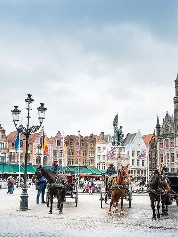Horse-drawn carriages and people in a historic European square with ornate buildings and cloudy sky.
