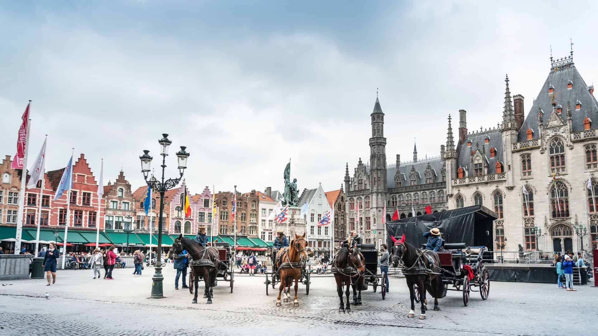 Horse-drawn carriages and people in a historic European square with ornate buildings and cloudy sky.