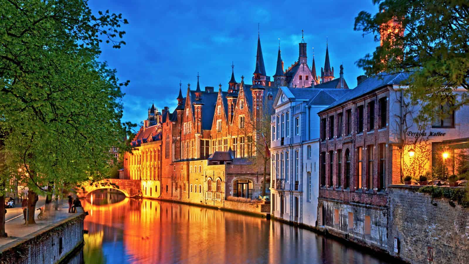 Historic buildings and trees line a glowing canal at dusk in Bruges, Belgium, with reflections in the water.