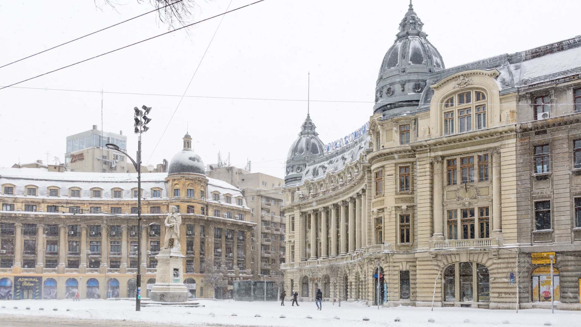 People walk in the snow near historic buildings and a statue in a city square during winter.