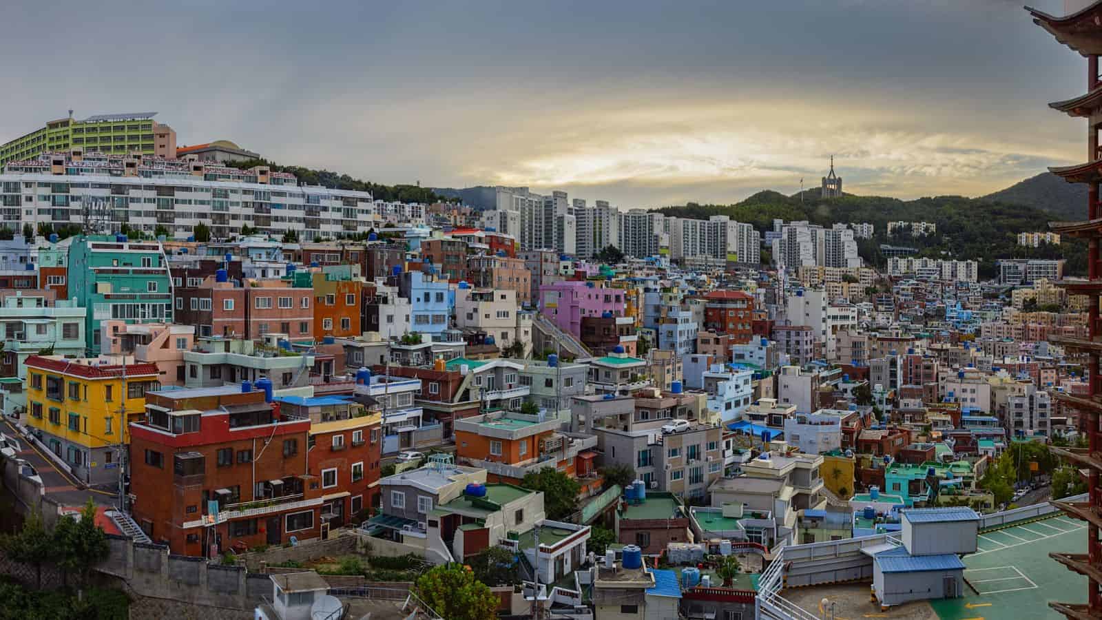 Colorful, densely packed buildings on a hillside with high-rises in the background under a cloudy sky.