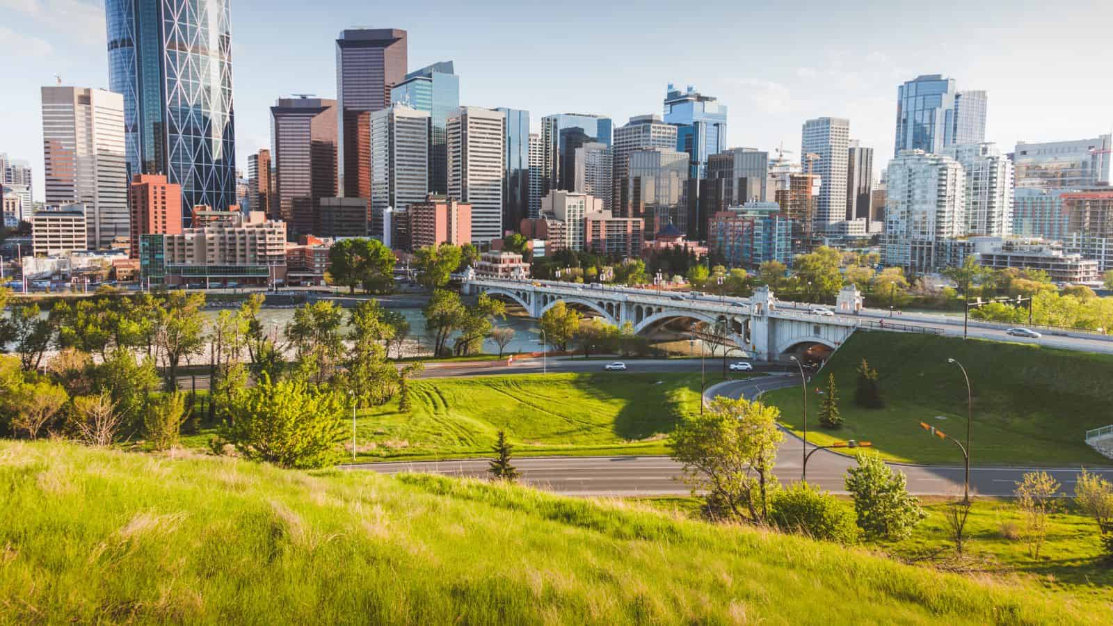 Downtown Calgary skyline with modern buildings, a river, and a bridge surrounded by green parkland.