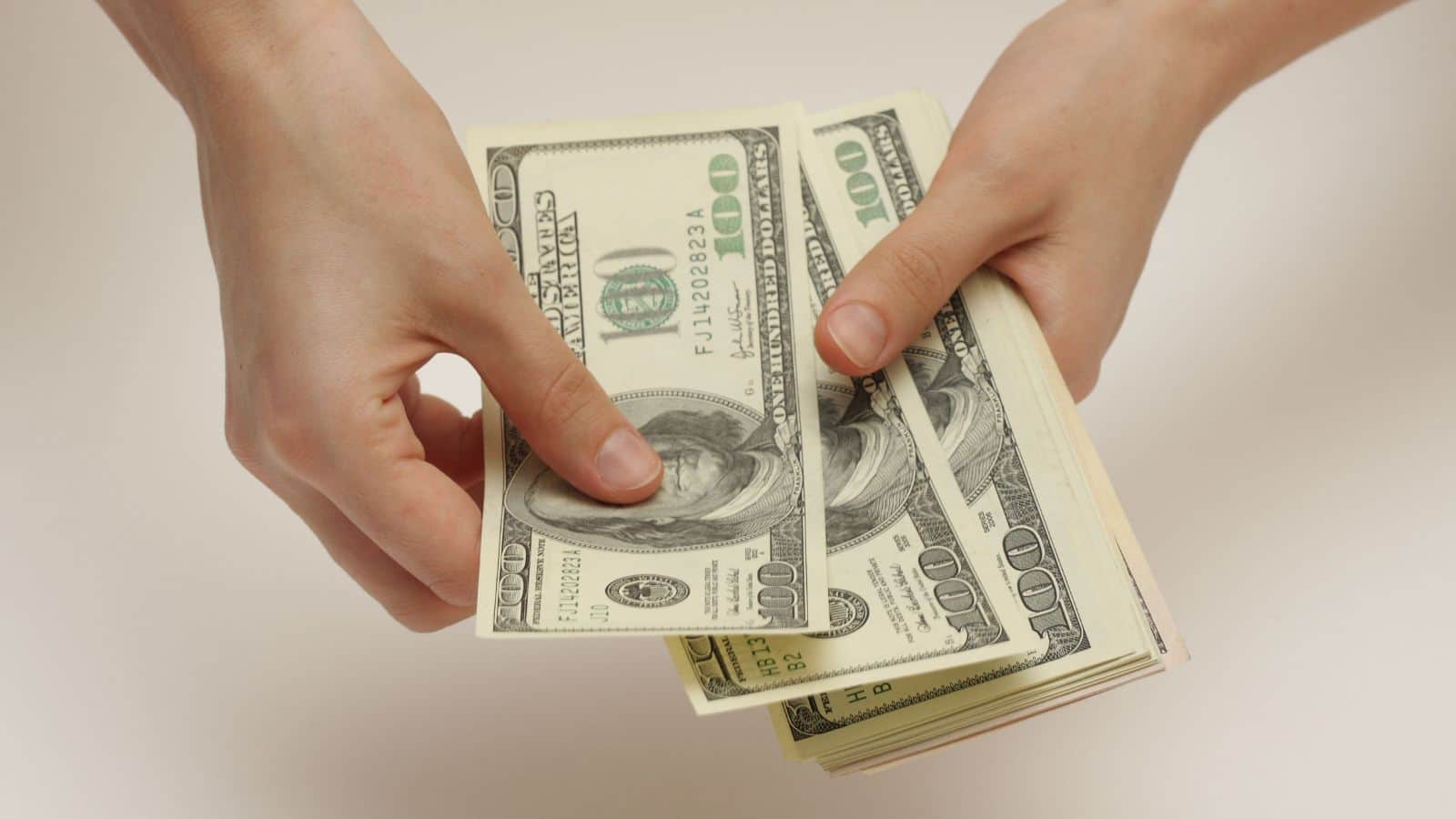A person hands over several $100 bills, holding a stack of cash against a plain light background.
