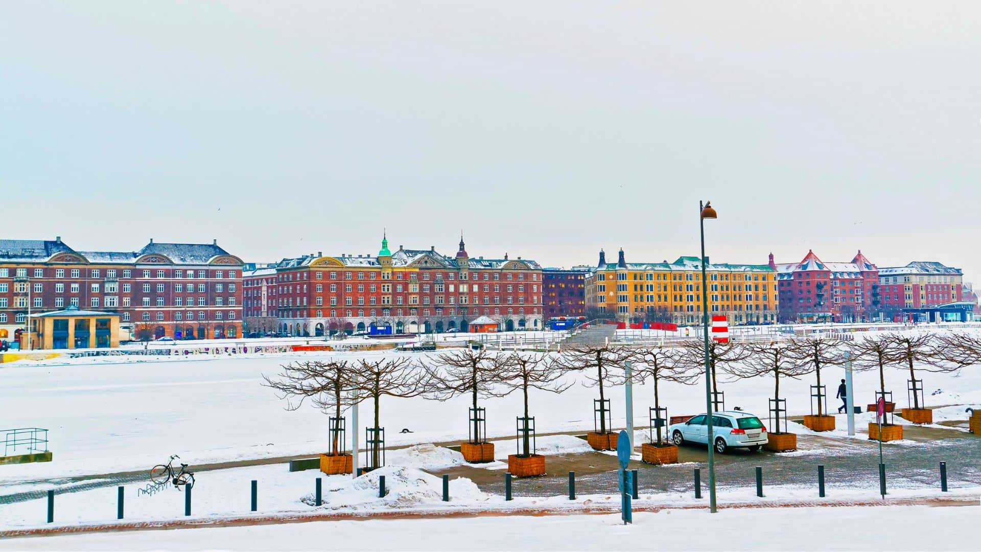 Snow-covered urban park with bare trees, parked cars, and colorful buildings in the background under a cloudy sky.