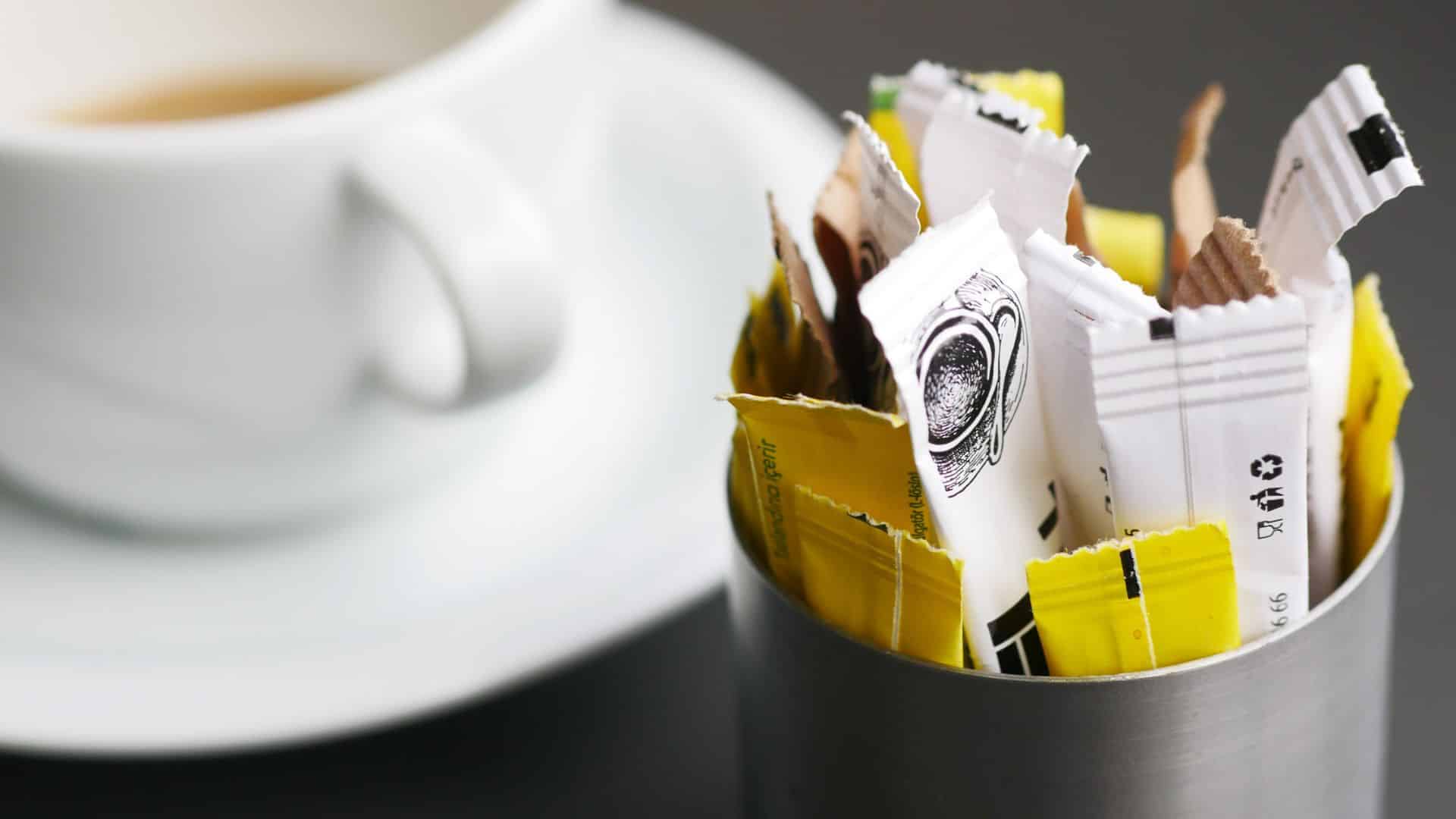 Packets of sugar or sweetener in a metal holder next to a white cup of coffee on a saucer.