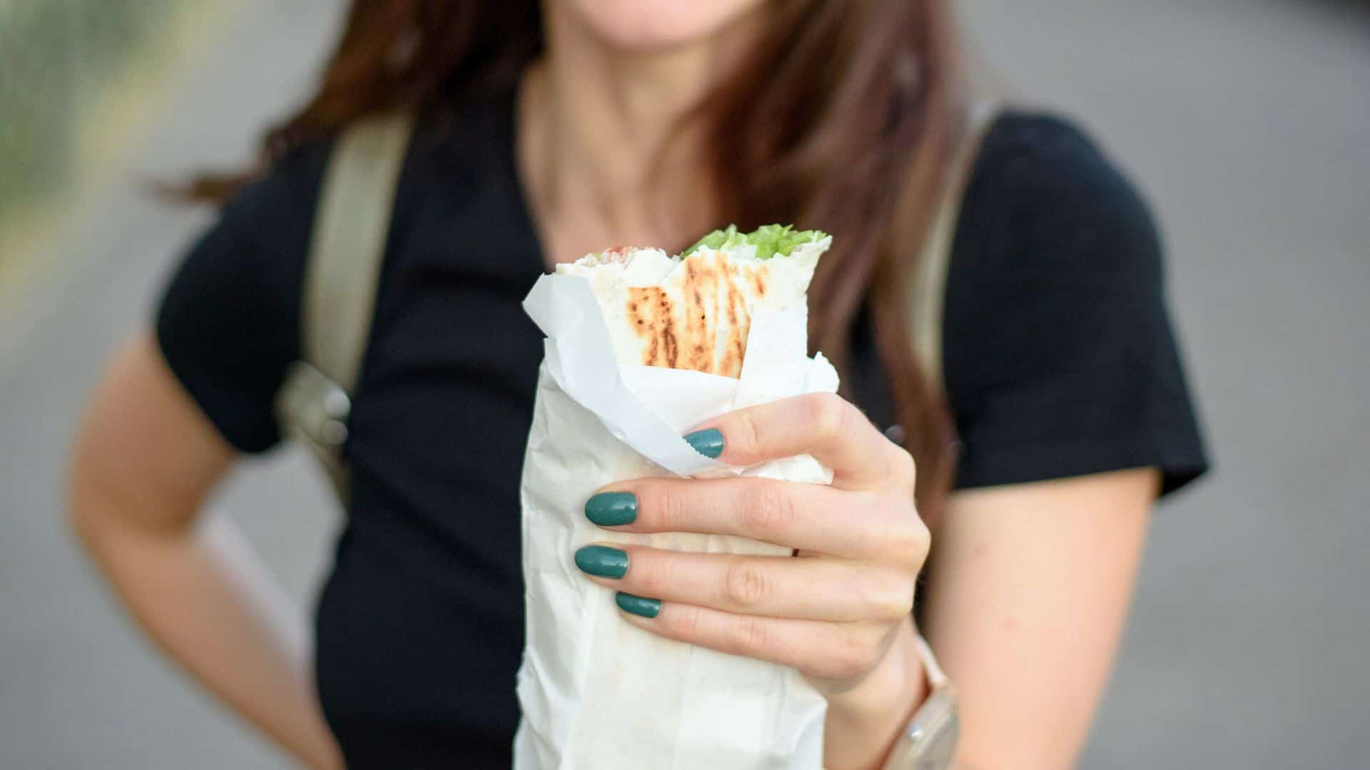 A woman holding a wrapped sandwich or wrap, partially eaten, outdoors, with green-painted nails.