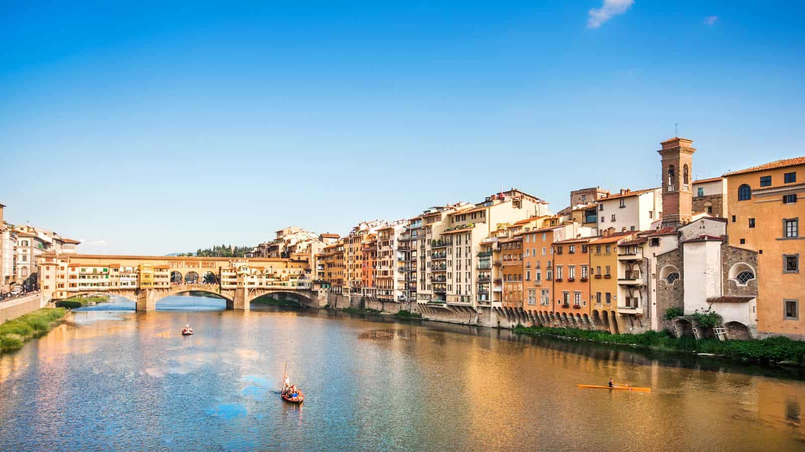 Colorful buildings and boats along the Arno River with Ponte Vecchio bridge in Florence, Italy, under a blue sky.