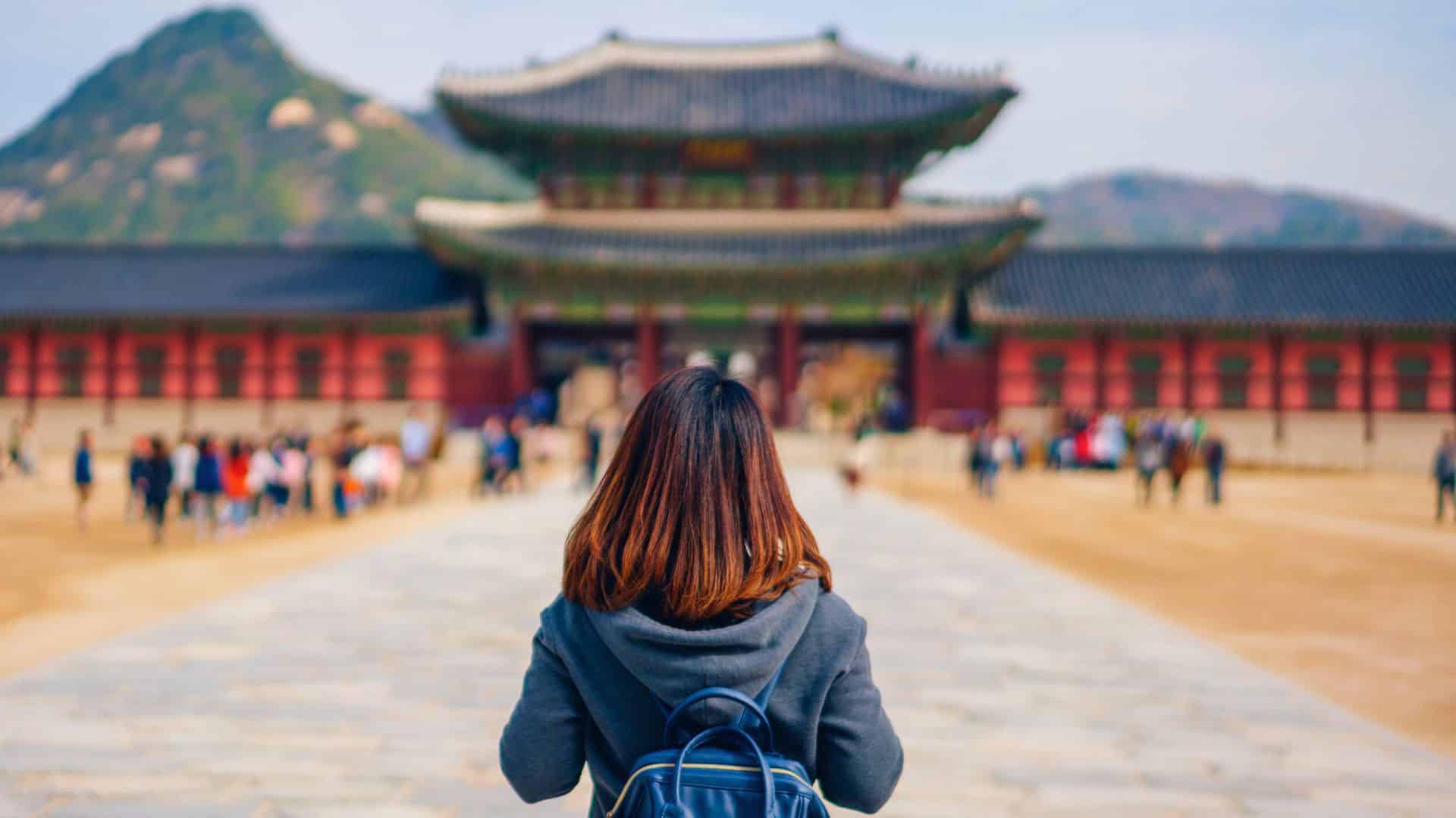 A woman with a backpack stands facing a traditional palace with mountains in the background.