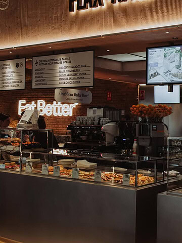 Bakery counter with pastries, cakes, and bread on display—including gluten free options—digital menu screens, and an "Eat Better" sign. Discover delicious treats at this Gluten Free Barcelona spot.