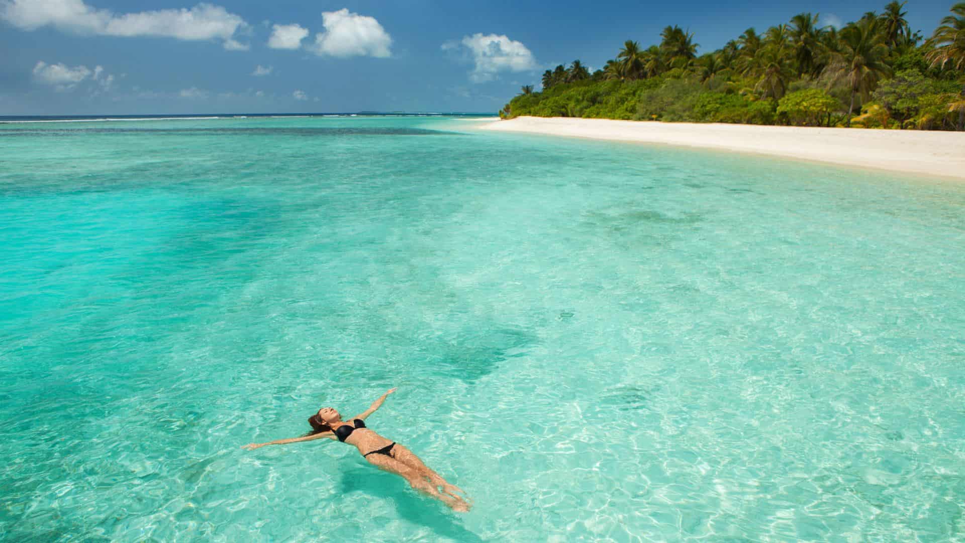 Woman in a black bikini floats in clear turquoise water near a tropical beach with palm trees.