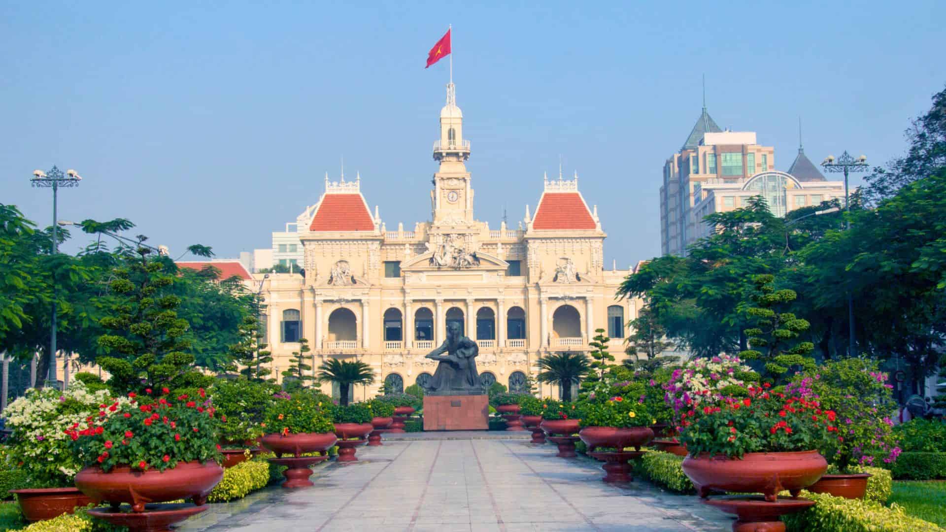 French colonial building with red roof, Vietnamese flag, and gardens in Ho Chi Minh City, Vietnam.