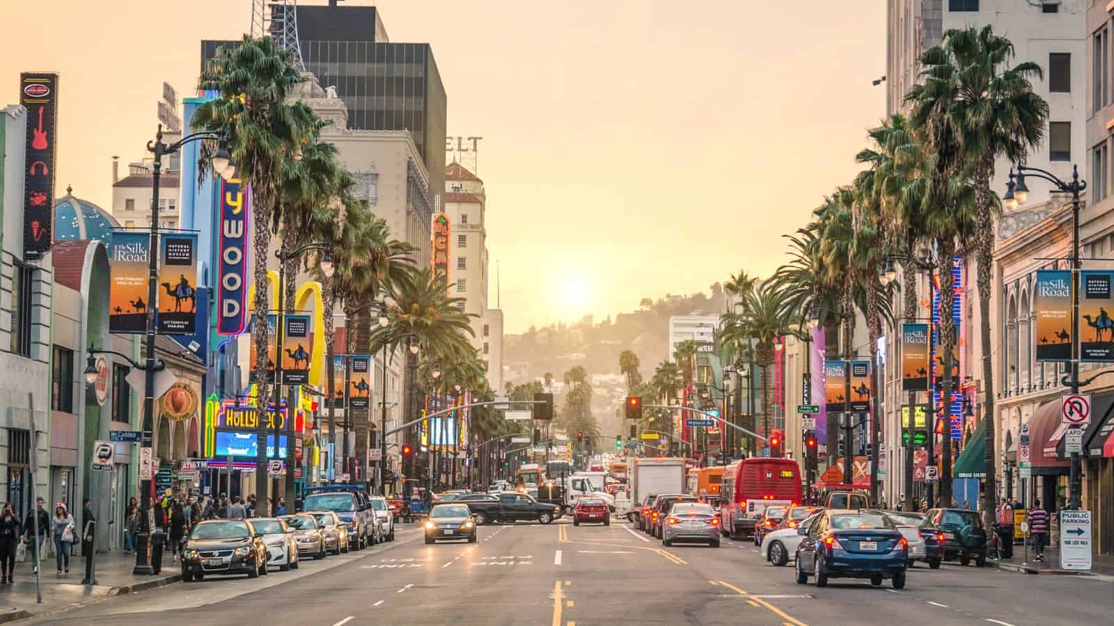 Busy Hollywood Boulevard at sunset, lined with palm trees, cars, and colorful signs and billboards.