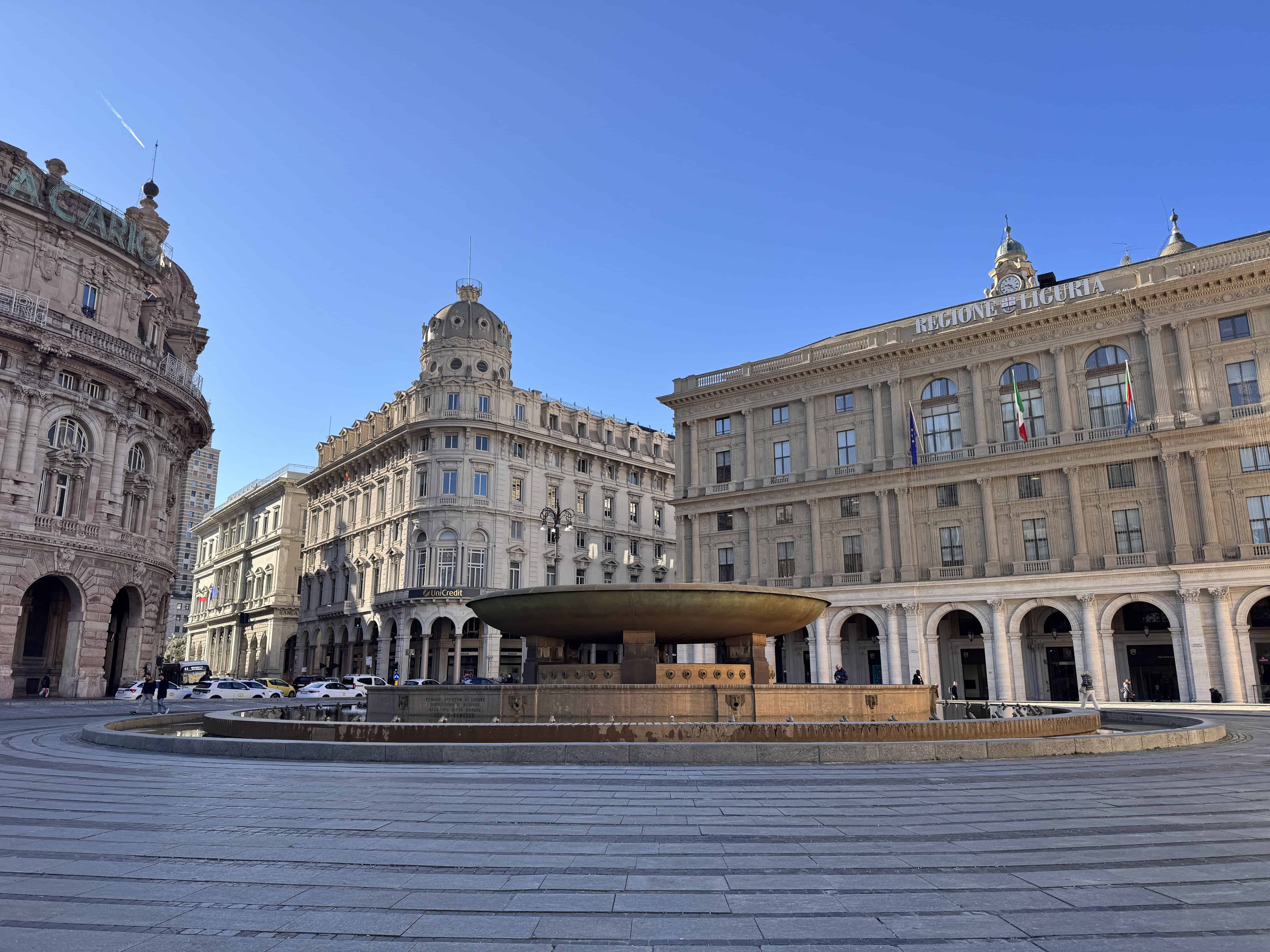 Large round fountain in a plaza surrounded by historic buildings under a clear blue sky in Genoa, Italy—a perfect spot to explore if you have just 24 hours in the city.