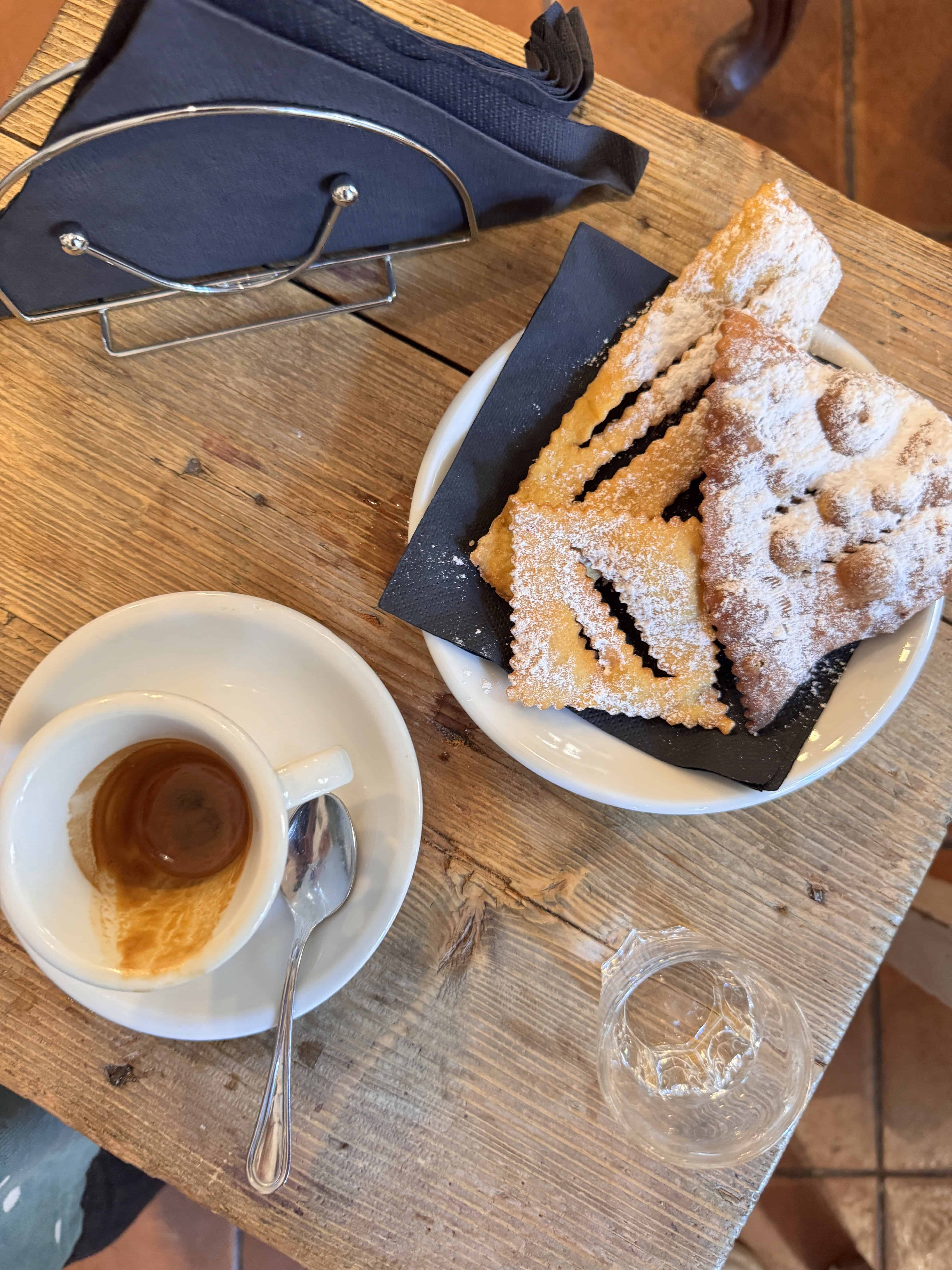 An empty espresso cup, glass of water, and sugar-dusted pastries rest on a rustic wooden table—capturing the essence of 24 hours in Genoa, Italy.