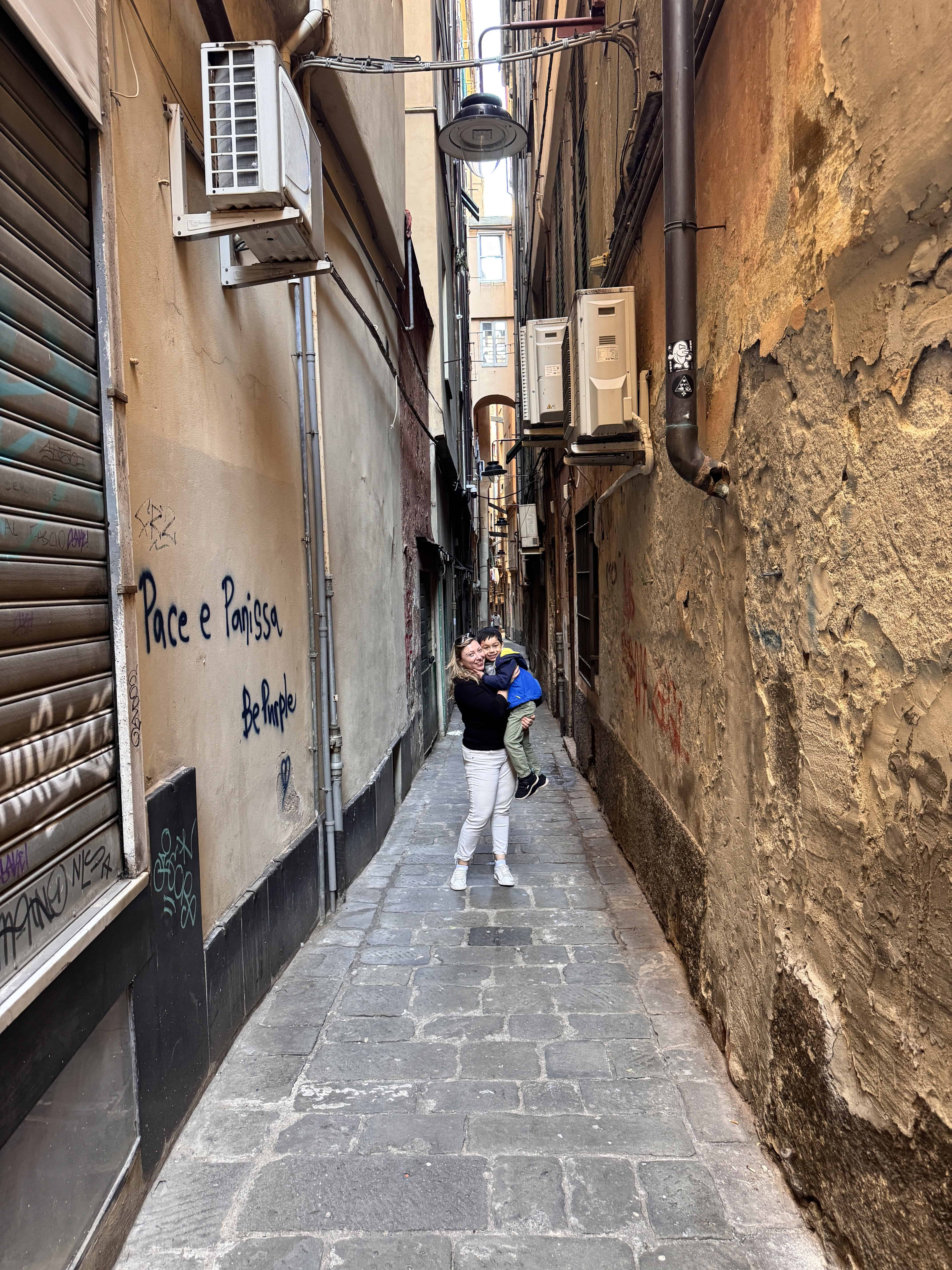 A person holds a child while standing in a narrow, graffiti-covered alleyway with stone pavement in Genoa, Italy, capturing the city’s unique character at any hour of the day.