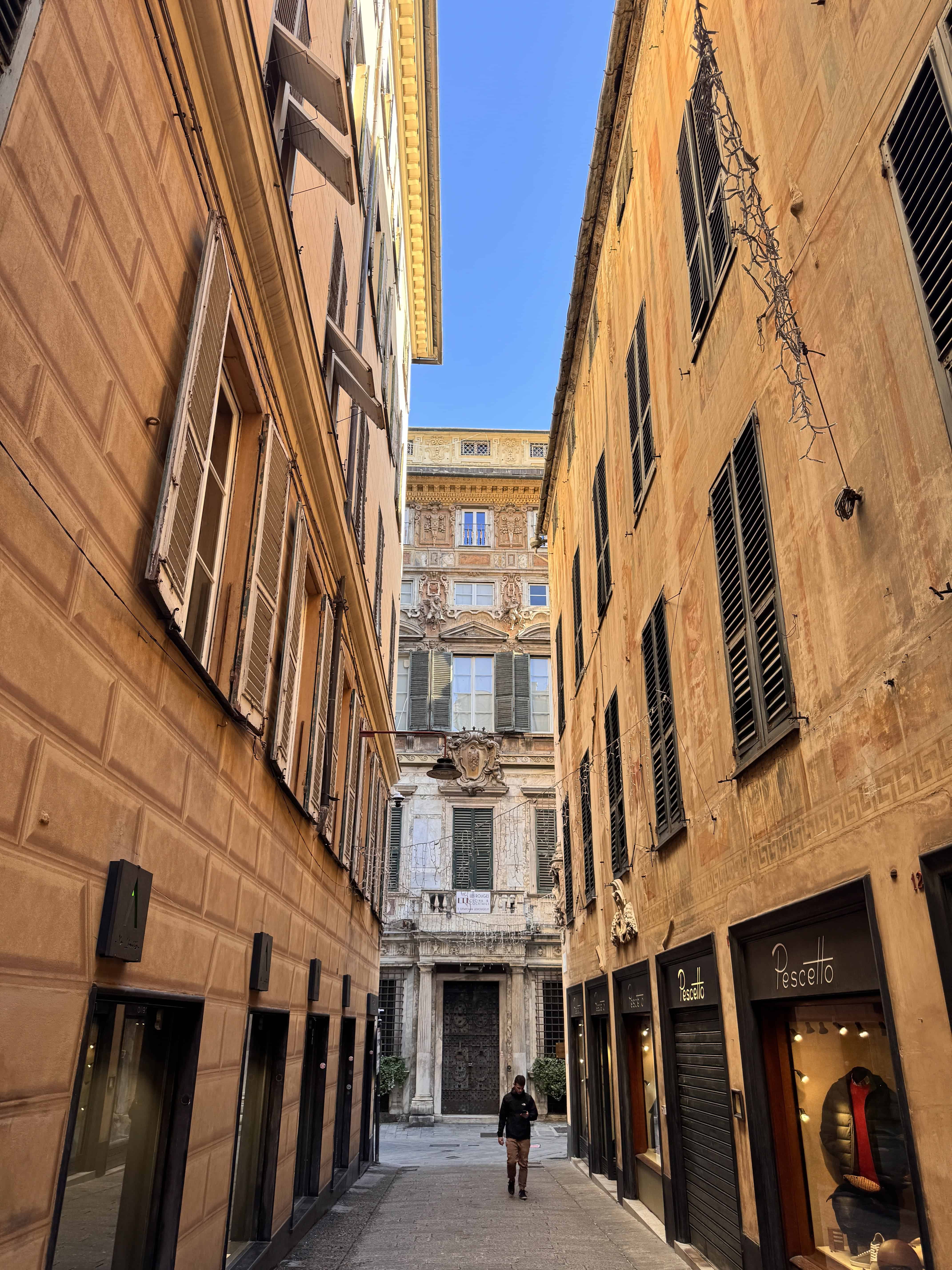 A narrow street in Genoa, Italy, with tall buildings, open windows, and a person walking toward an ornate doorway—capturing city life at any hour of the day.
